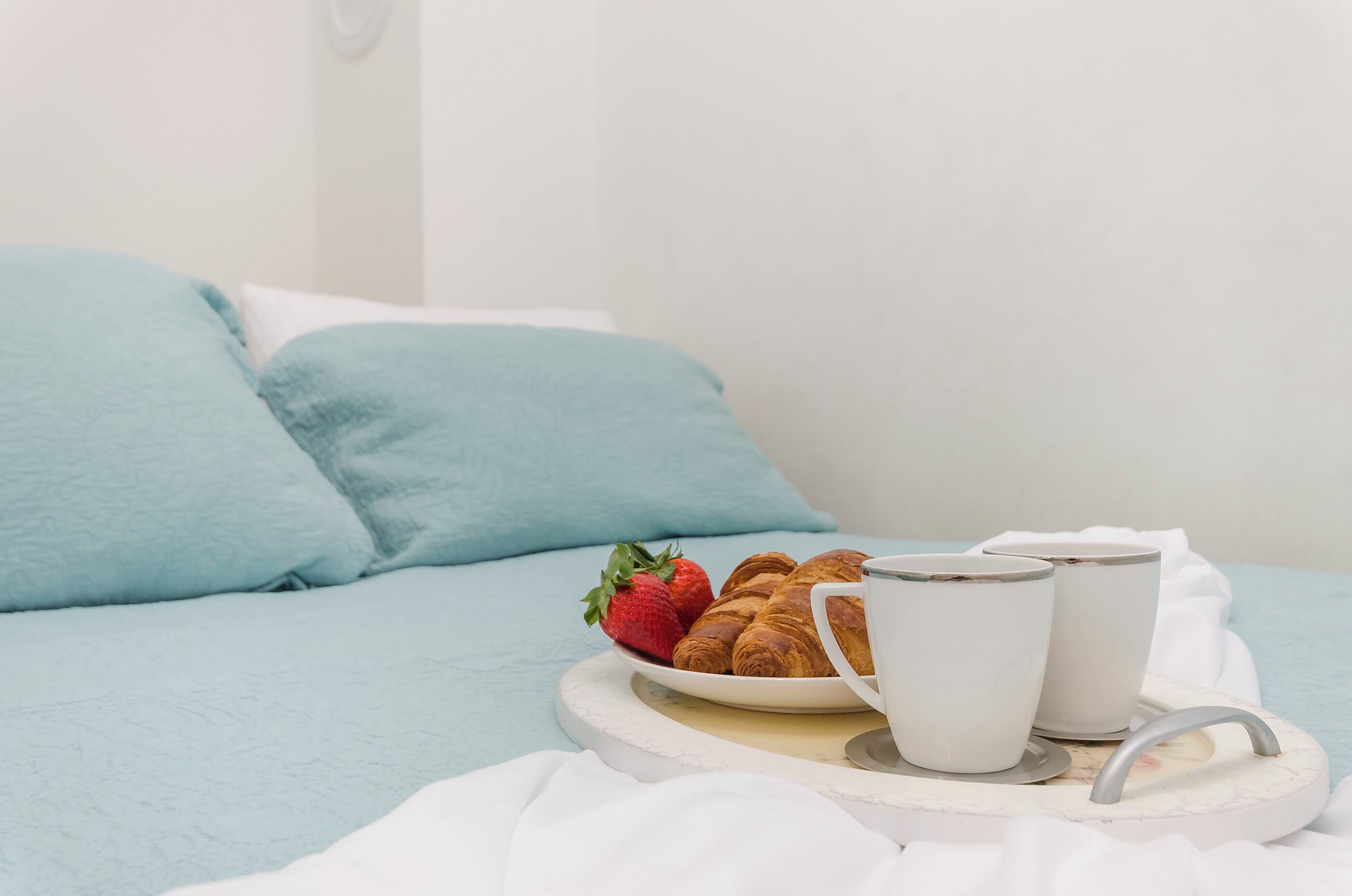 A bed with light blue bedding, a tray with croissants, a strawberry, and two white mugs.