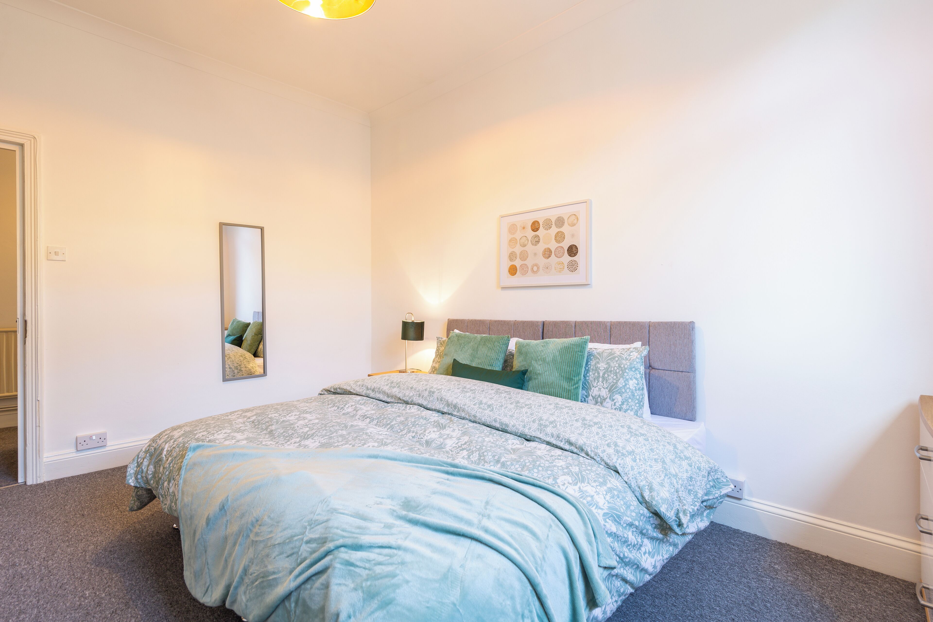 Quarto com cama, mesa de cabeceira, espelho fixado na parede e quadro emoldurado.