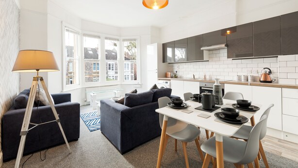 A modern kitchen with white cabinets, a dining table with chairs, and a large window providing a view of the city. - Stunning 5-bed Maisonette in Bristol (Bristol)