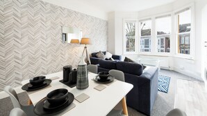 A modern living room with a dining area, featuring a herringbone-patterned wall, a dark blue sofa, and a glass coffee table. - Stunning 5-bed Maisonette in Bristol (Bristol)