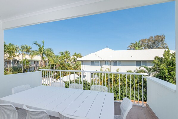 A balcony with a white table and chairs, overlooking a residential area with palm trees and houses.