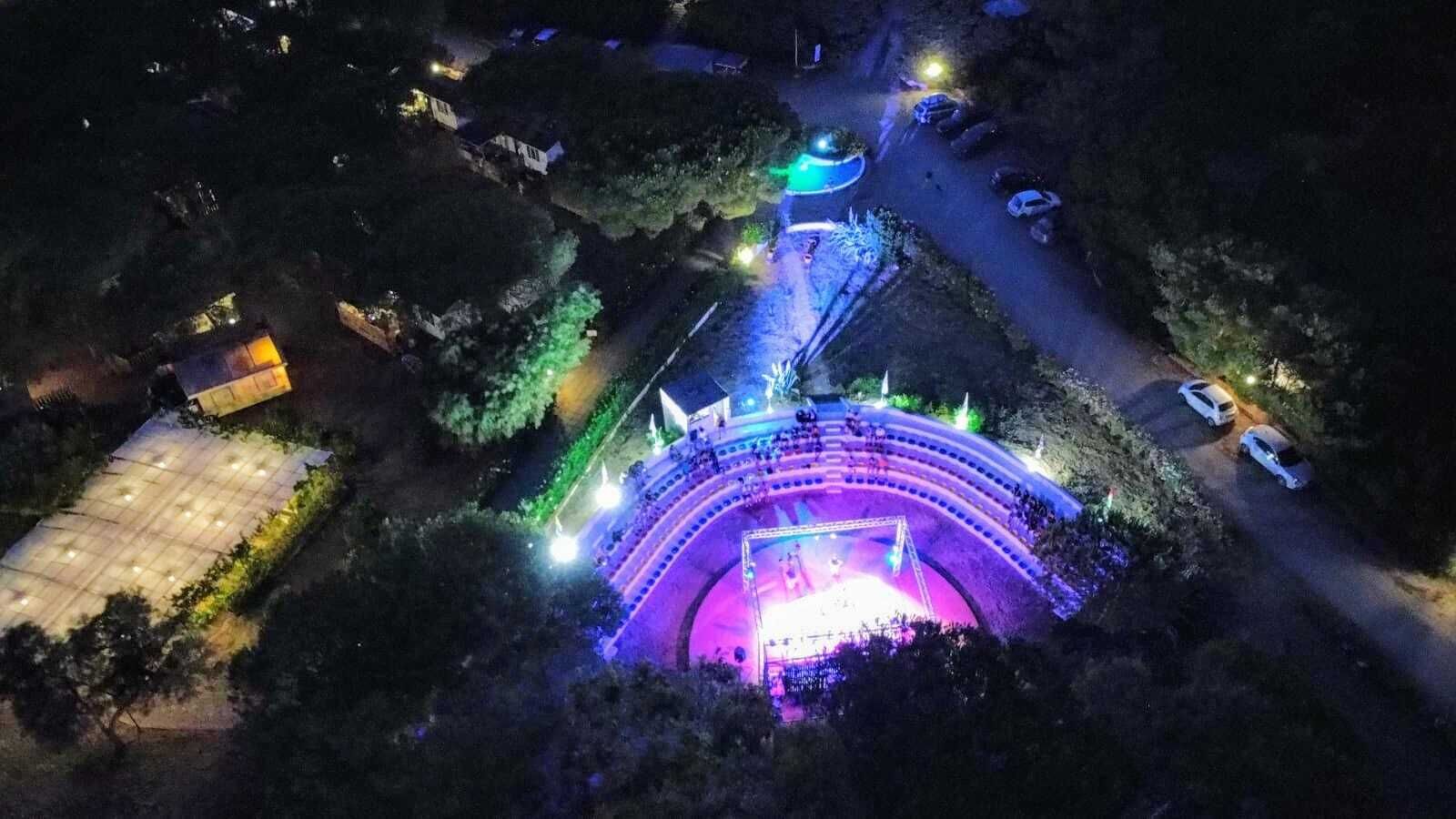 Vue aérienne d’un amphithéâtre circulaire doté de gradins et d’une scène centrale, entouré d’arbres et d’un espace de stationnement.