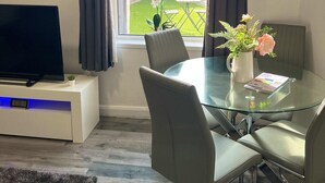 A modern living room with a glass dining table, grey chairs, a flat-screen TV on a white cabinet, and a view of a garden through a window with dark curtains. - Birmingham Blissful Retreat - 4 Bedroom (Birmingham)