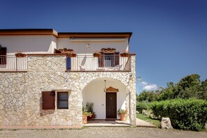 A two-story stone building with a balcony, a central archway, and potted plants. - Villa Parco an oasis of peacefulness in the heart of the Porto Conte Nature Park (Sardegna)