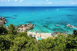 Une plage côtière aux eaux turquoise et limpides, parsemée de formations rocheuses, où les gens profitent du soleil, de la plage et de la mer.