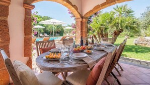 Un área de comedor al aire libre con una mesa de madera dispuesta para cuatro personas, rodeada de arcos de piedra, con una piscina y vegetación al fondo.
