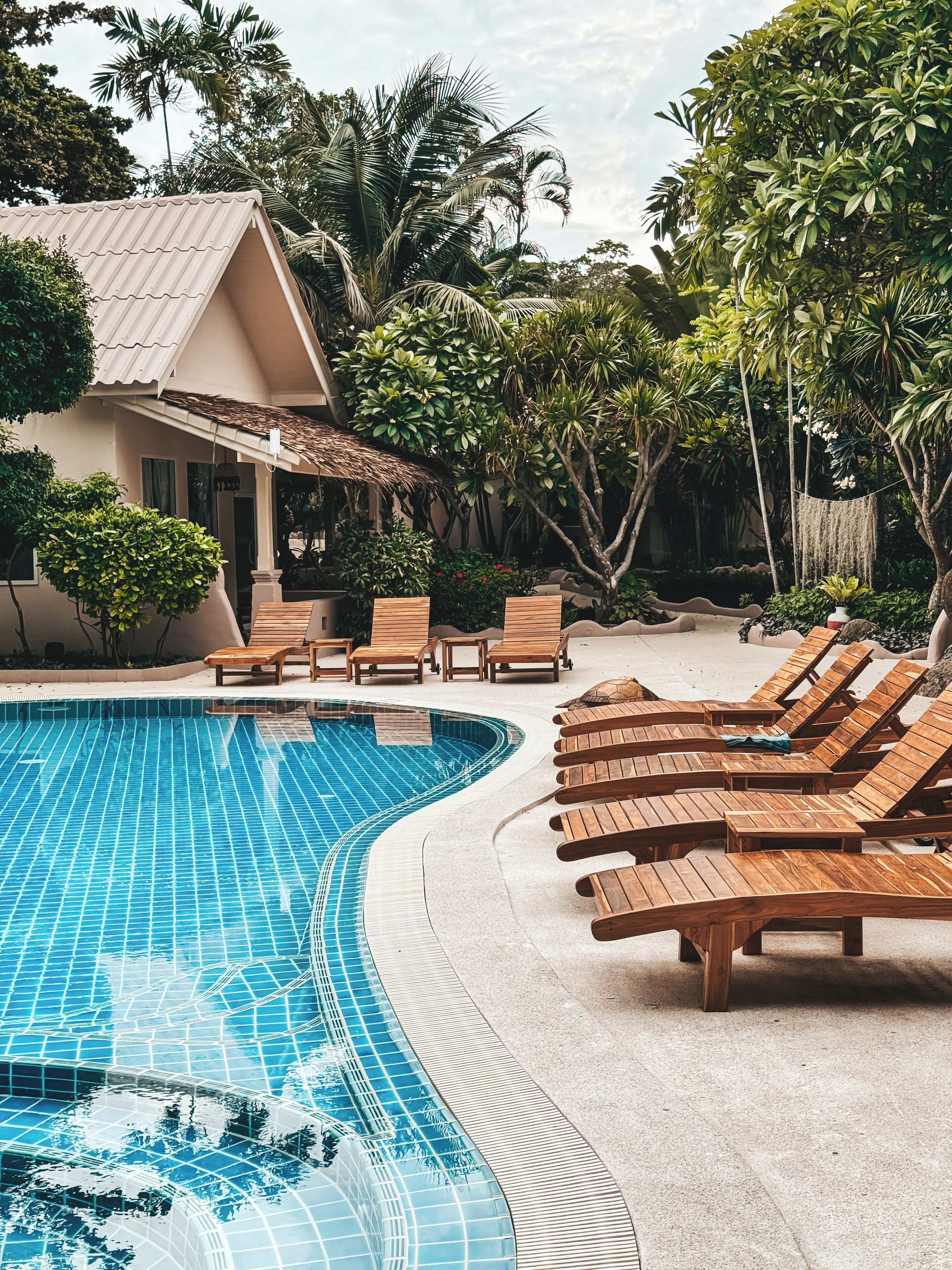 A pool area with a tiled swimming pool, wooden lounge chairs, and a house with a tiled roof.