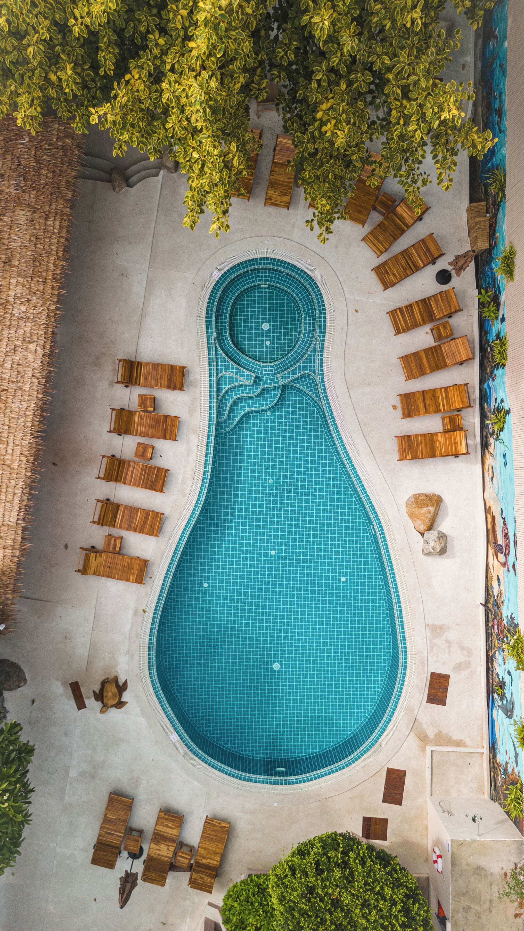Ein Pool mit ungewöhnlicher Form, umgeben von Holzliegen und einem Wandgemälde.