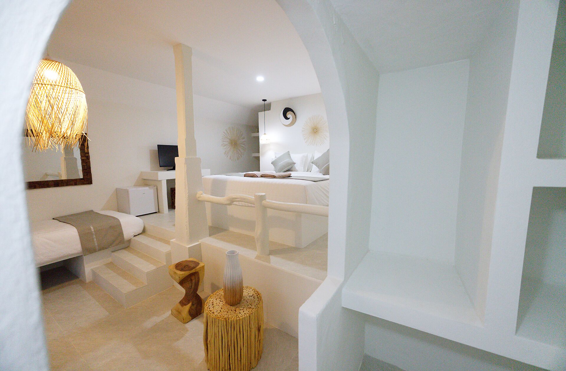 A modern, minimalist hotel room with a white color scheme, a large mirror, a television, a bed with white bedding, and a unique wooden stool.