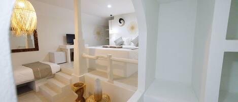 A modern, minimalist hotel room with a white color scheme, a large mirror, a television, a bed with white bedding, and a unique wooden stool.