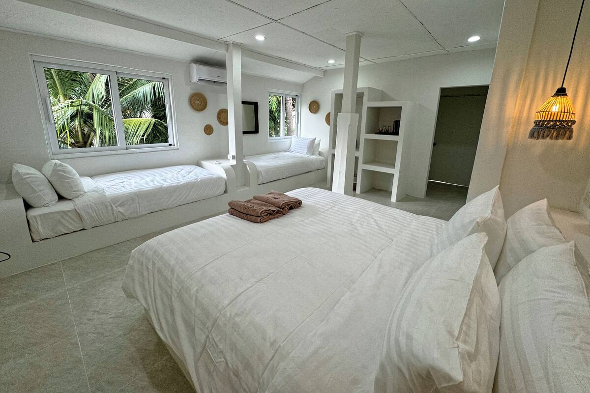 A modern hotel room with a large bed, two smaller beds, a built-in shelf, and a view of palm trees through the windows.