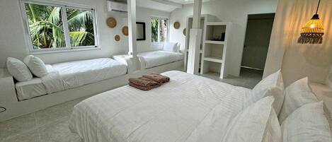 A modern hotel room with a large bed, two smaller beds, a built-in shelf, and a view of palm trees through the windows.