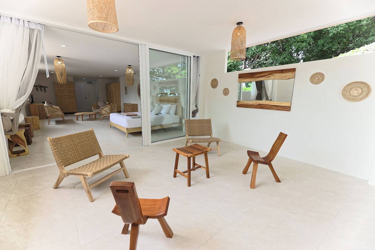 A spacious room with a large bed, a sofa, and two wooden chairs. There is a small table and a hanging light fixture.