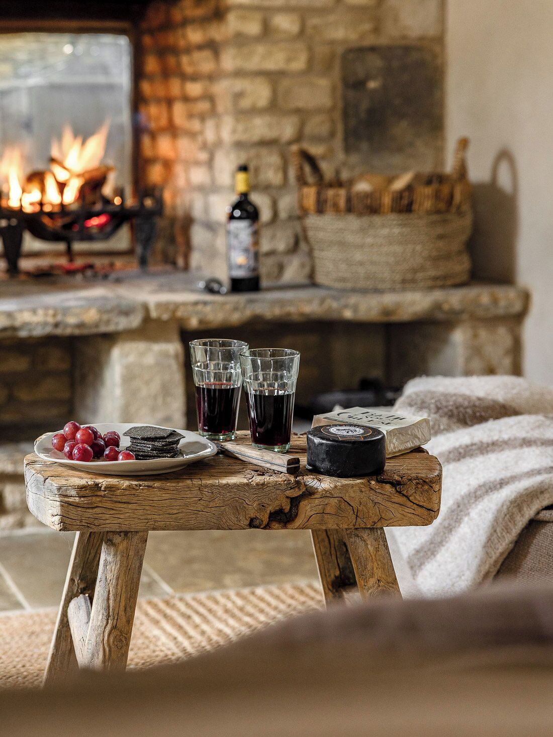 A rustic wooden table with a plate of grapes, two glasses of red wine, and a cheese wheel. A fireplace with a bottle of wine and a basket is in the background.