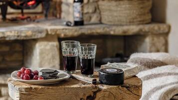 A rustic wooden table with a plate of grapes, two glasses of red wine, and a cheese wheel. A fireplace with a bottle of wine and a basket is in the background.