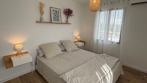 Ein Schlafzimmer mit einem Bett, Nachttischlampen, einer Pendelleuchte, einem Fenster mit durchscheinenden Vorhängen und Wandregalen mit Dekoration.