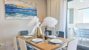 Un comedor con una mesa de madera dispuesta para seis personas, sillas blancas, un jarrón decorativo con plumas blancas y un cuadro en la pared.