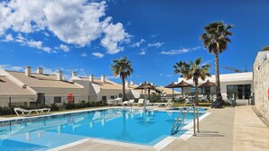 Una piscina con sillas de descanso y palmeras, rodeada de edificios con techos de tejas.