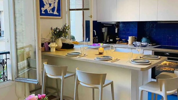 A modern kitchen with white cabinets, a central island with bar stools, a dining area with a table and chairs, and a large window with blinds. - Chic 3 Pièces Familial Pour Bébé au Cœur de Paris (Paris)