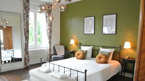 Ein ordentlich eingerichtetes Schlafzimmer mit einem großen Bett, zwei Nachttischen mit Lampen, einem Sessel, einem Spiegel und einem Fenster mit Vorhängen.