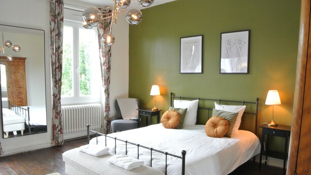 A neatly arranged bedroom with a large bed, two bedside tables with lamps, a chair, a mirror, and a window with curtains.