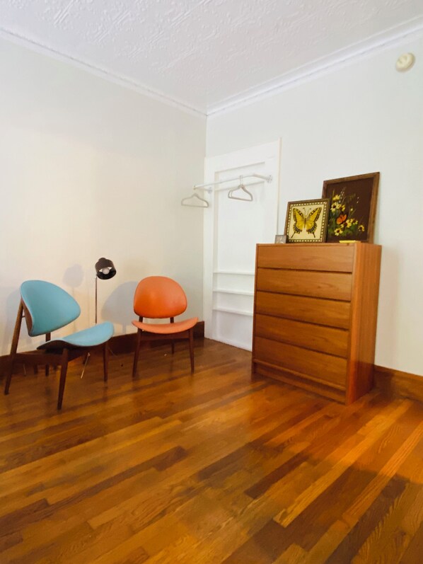 Una habitación con suelo de madera, una silla azul y una naranja, un aparador de madera y un perchero de pared con dos perchas.