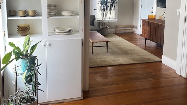 A spacious living room with a wooden floor, a white cabinet with kitchenware, a grey sofa, and a large window with blinds.