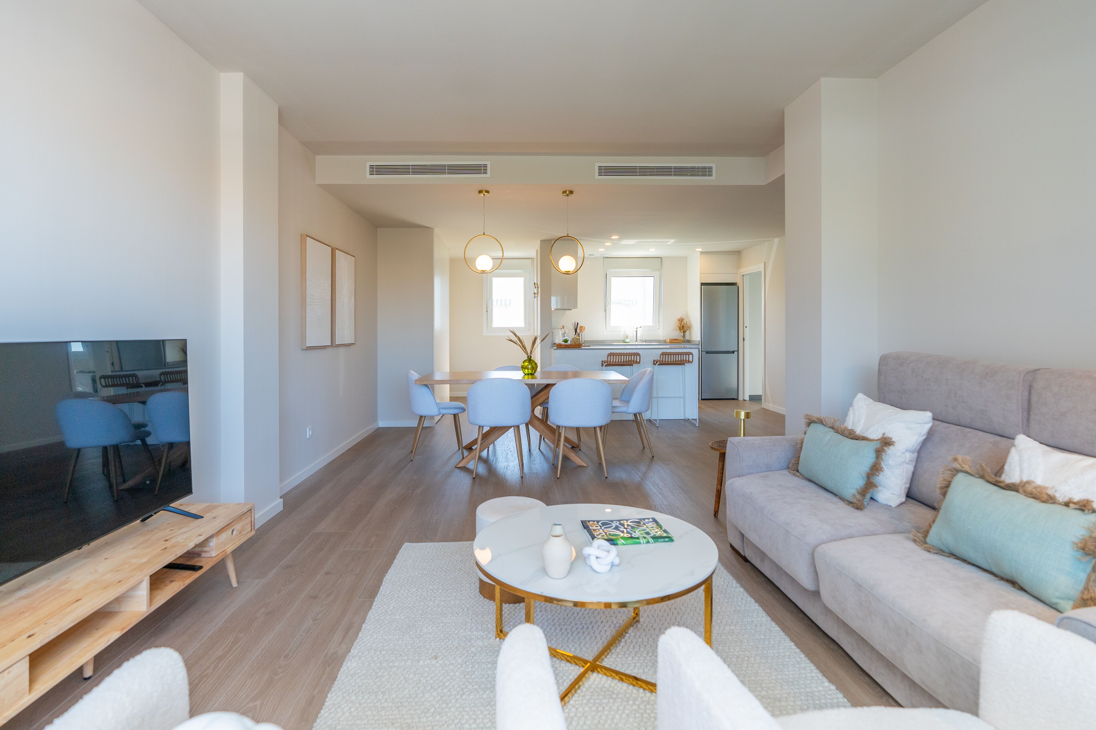 Ein modernes Wohnzimmer mit Sofa, Couchtisch und TV-Schrank. Ein Essbereich mit Stühlen und Tisch ist ebenfalls vorhanden, und im Hintergrund befindet sich eine Küche.