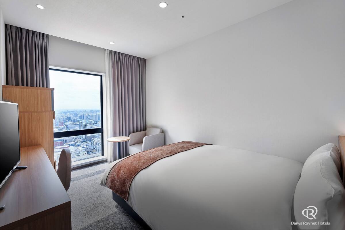 Una habitación de hotel moderna con una cama grande, un escritorio con una computadora, una silla y vistas a la ciudad a través de un ventanal amplio.