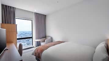 A modern hotel room with a large bed, a desk with a computer, a chair, and a view of the city through a large window.