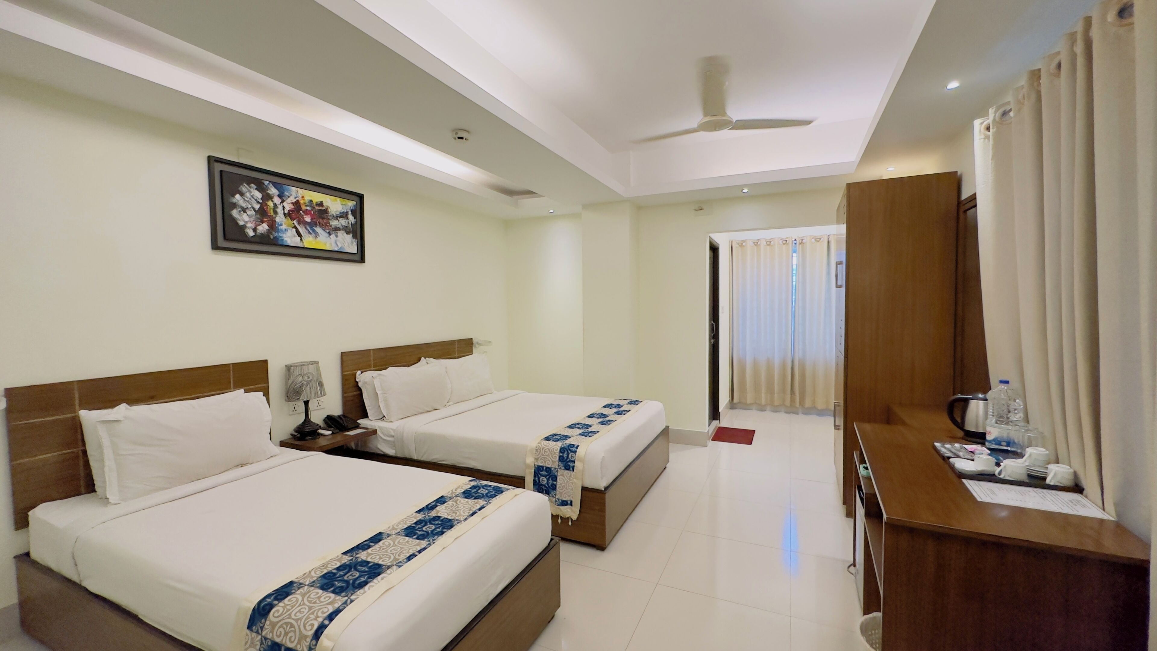 A hotel room with two beds, a desk with a chair, a ceiling fan, and a painting on the wall.