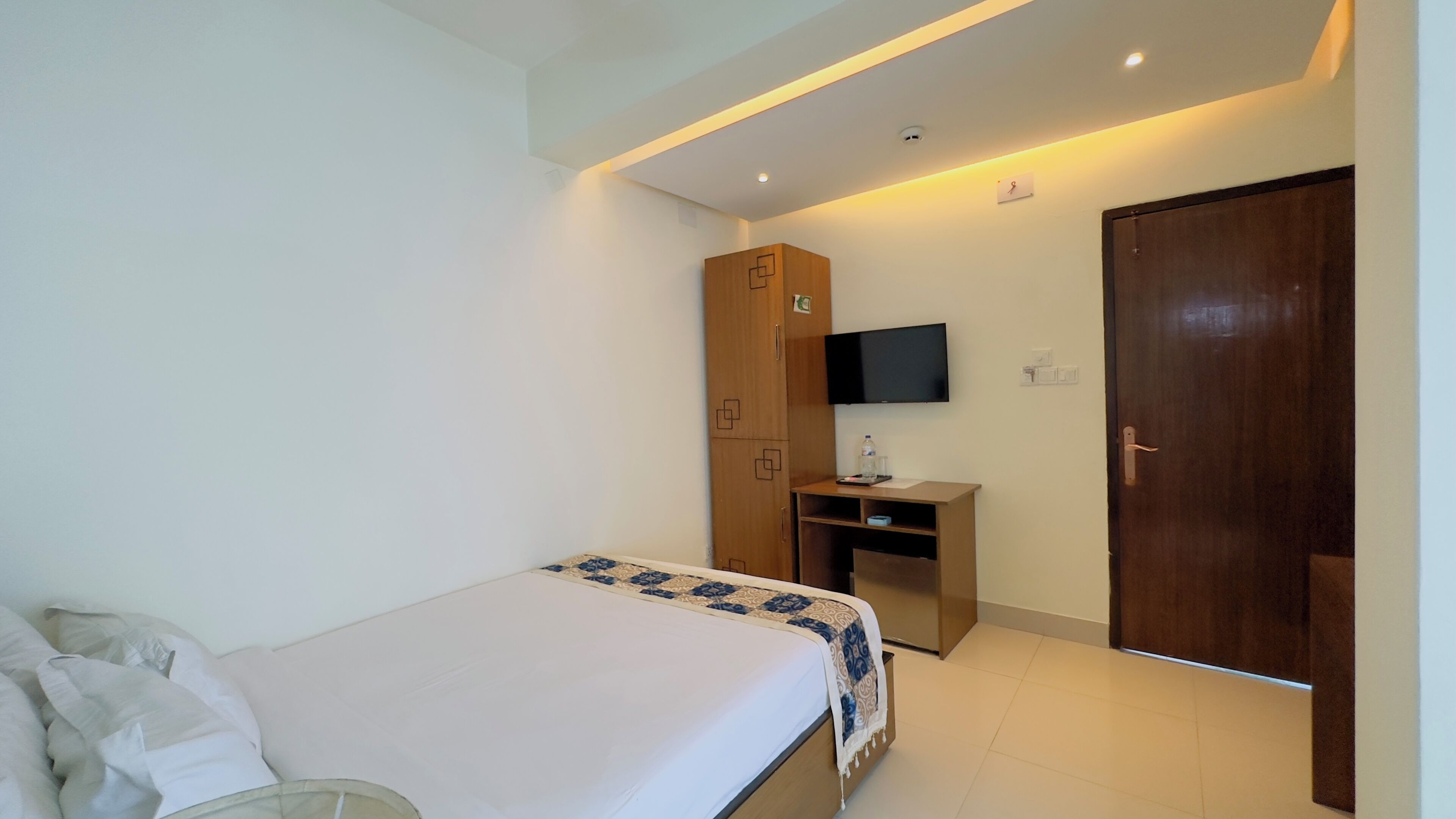 A bedroom with a bed, a wooden wardrobe, a desk with a flat-screen TV, and a door.