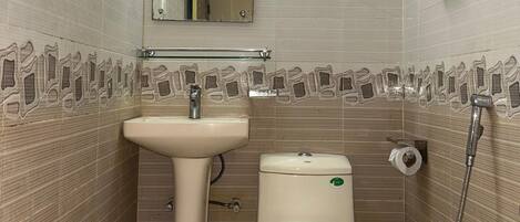 Une petite salle de bain avec un lavabo sur colonne, des toilettes et une douche. Un miroir est placé au-dessus du lavabo, et un porte-serviettes est fixé au mur.
