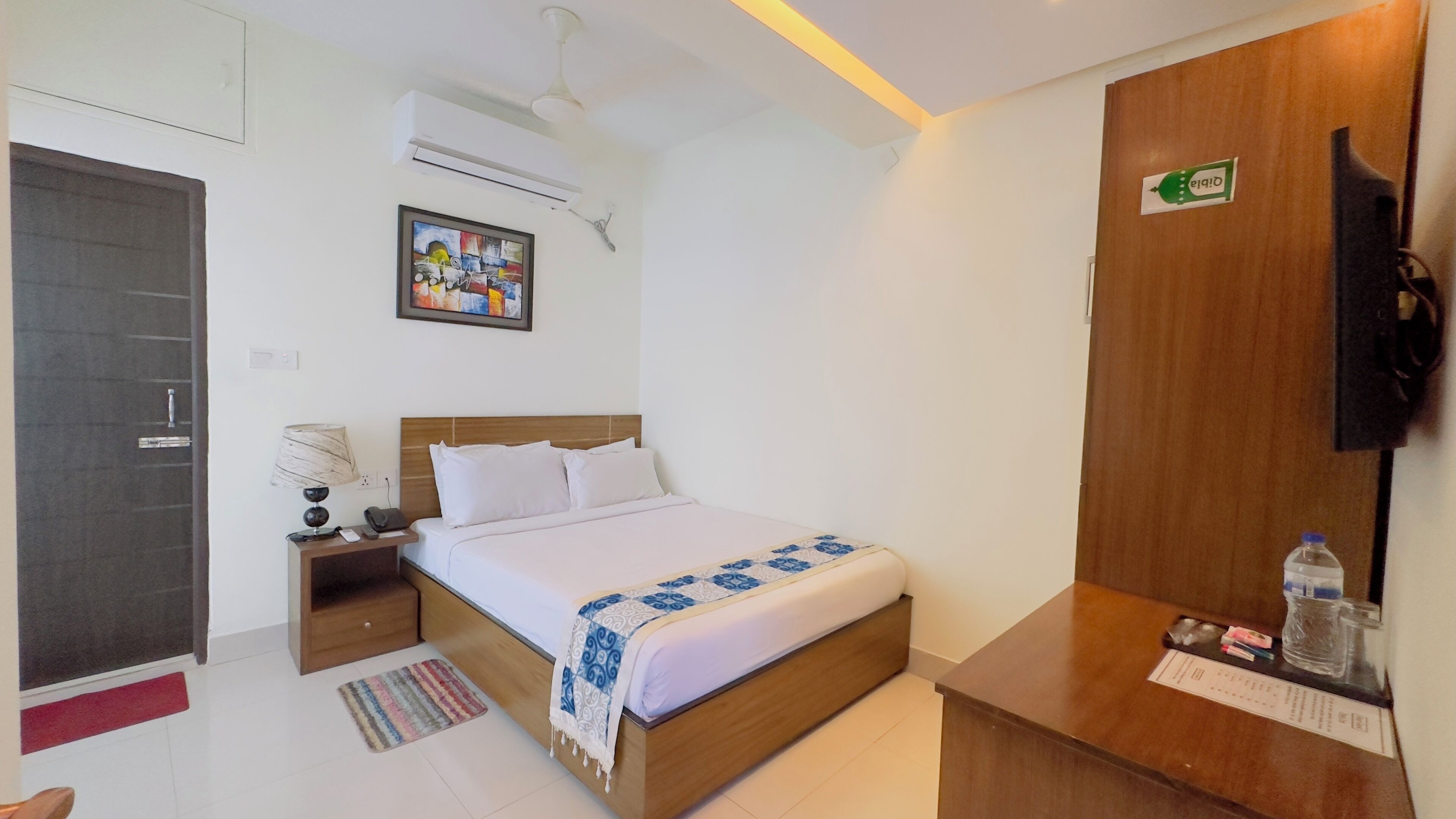 A hotel room with a bed, a nightstand, a desk, a chair, a wall-mounted TV, an air conditioning unit, and a door.