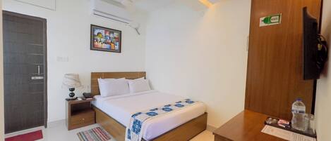 A hotel room with a bed, a nightstand, a desk, a chair, a wall-mounted TV, an air conditioning unit, and a door.