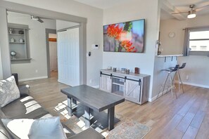 Ein modernes Wohnzimmer mit einer Couch, einem Couchtisch und einem Essbereich mit einer Bank. Es gibt eine Küchenzeile mit einer Bar und Hockern.