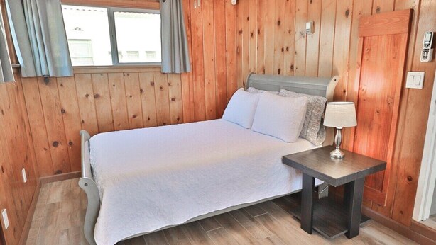 A wooden-paneled room with a bed, a bedside table with a lamp, and a window with curtains. - Campus Casita close to U of A and shopping (Tucson)