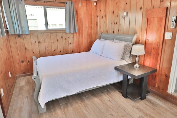 A wooden-paneled room with a bed, a bedside table with a lamp, and a window with curtains. - Campus Casita close to U of A and shopping (Tucson)