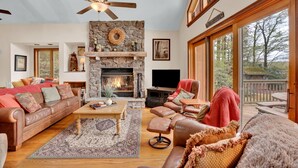 A spacious living room with a stone fireplace, leather furniture, and a large window overlooking a deck. - Lakefront | Private Dock | Family Friendly | Kayaks | Hot Tub | Row Boat | Fireplace (Lake Ariel)