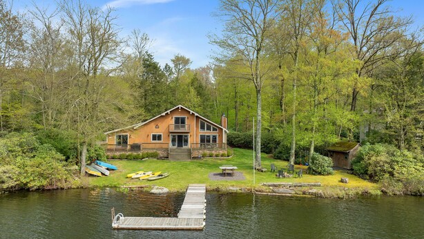 A cabin with a dock extending into a calm lake, surrounded by lush greenery and trees. - Lakefront | Private Dock | Family Friendly | Kayaks | Hot Tub | Row Boat | Fireplace (Lake Ariel)