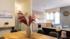 A modern kitchen with white cabinets, a dining area with a wooden table, and a living area with a sofa and a clock on the wall.