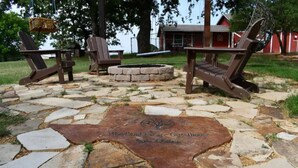 A stone patio with Adirondack chairs and a fire pit, surrounded by a wooded area.
