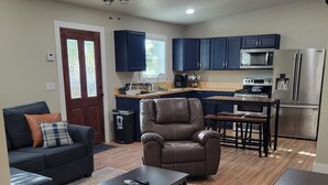 A modern living room with a sofa, armchair, coffee table, and a dining area with a table and chairs. A kitchen with dark cabinets and appliances is visible in the background.