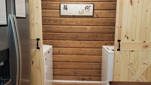 A compact kitchen with wooden panel walls, a stainless steel refrigerator, a white washing machine, and a wooden table.