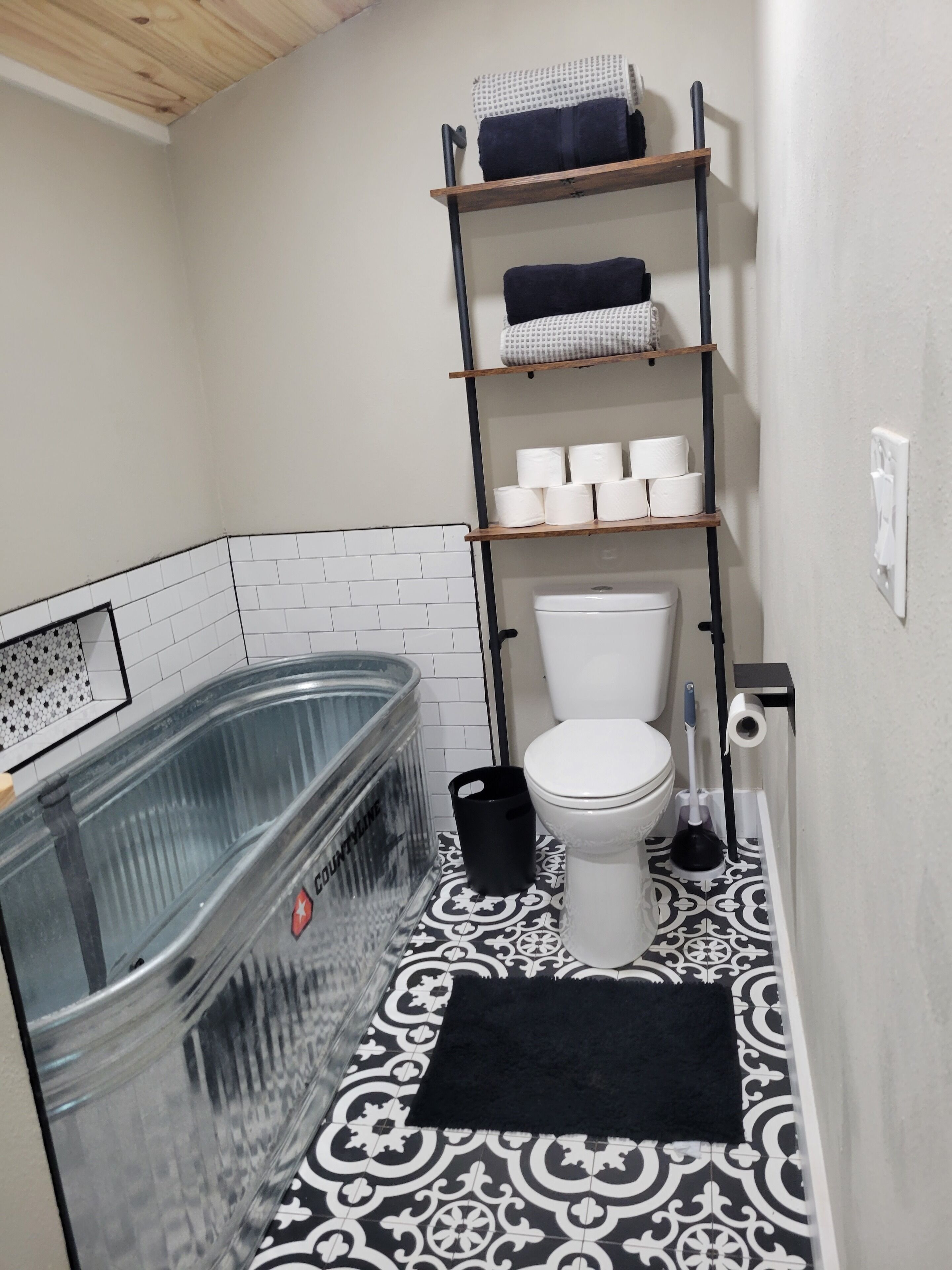 A bathroom with a glass bathtub, a toilet, a shelf with towels, and a black rug.