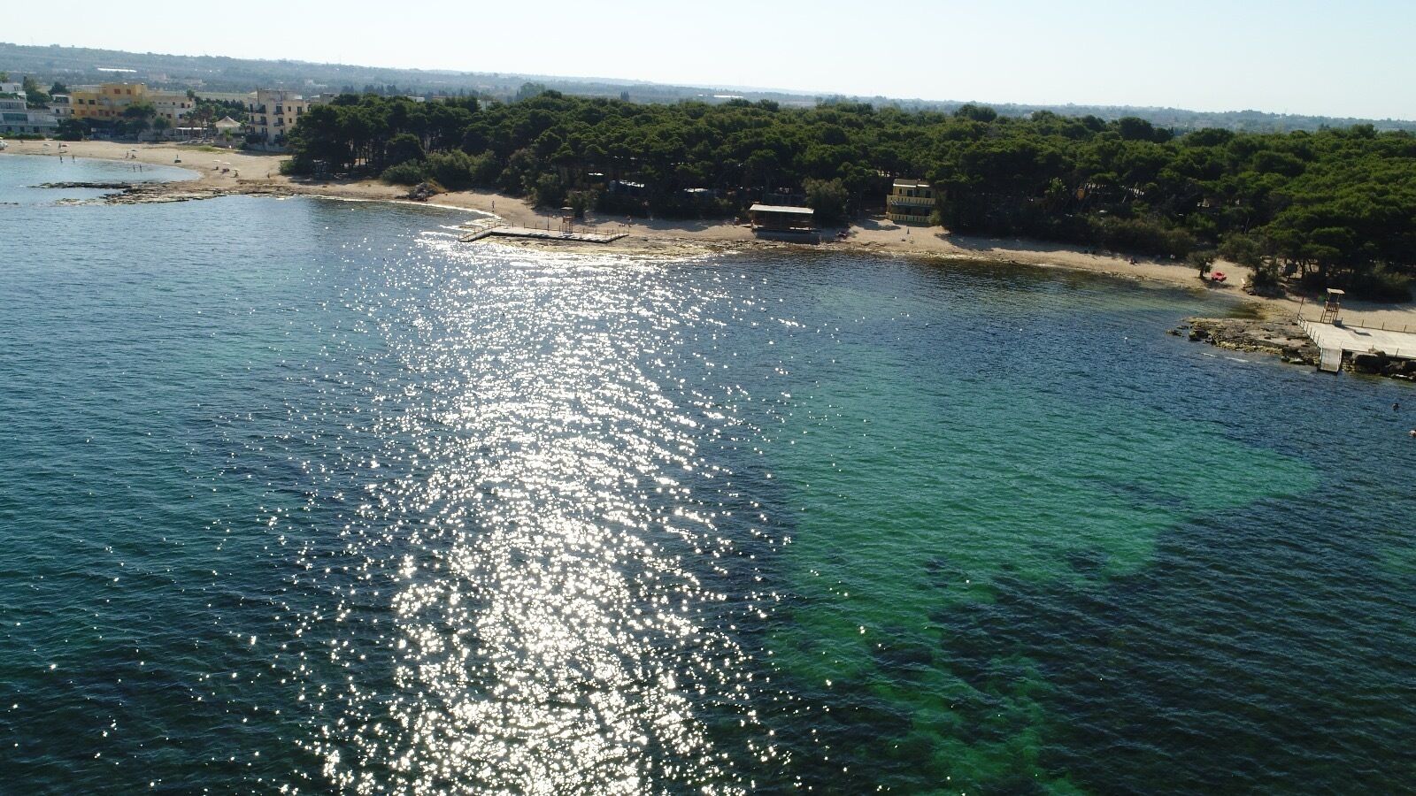 Et kystområde med klart vand, der spejler sollys, en sandstrand og bygninger langs kysten.