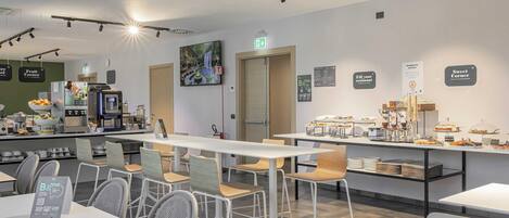 Un café moderno con mesas y sillas blancas, una barra con exhibición de alimentos y una pared verde con frutero.