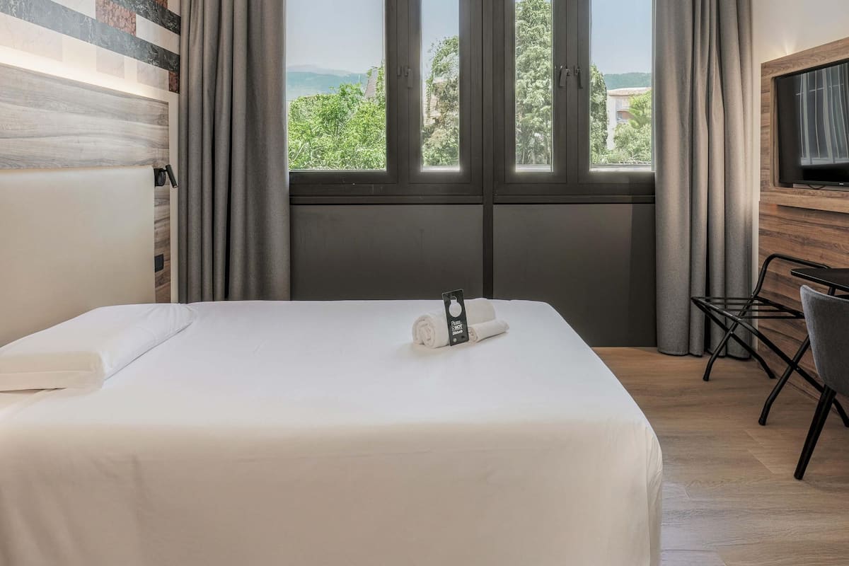 Une chambre d’hôtel avec un grand lit, un bureau avec une chaise, une télévision et une vue sur la verdure par les fenêtres.
