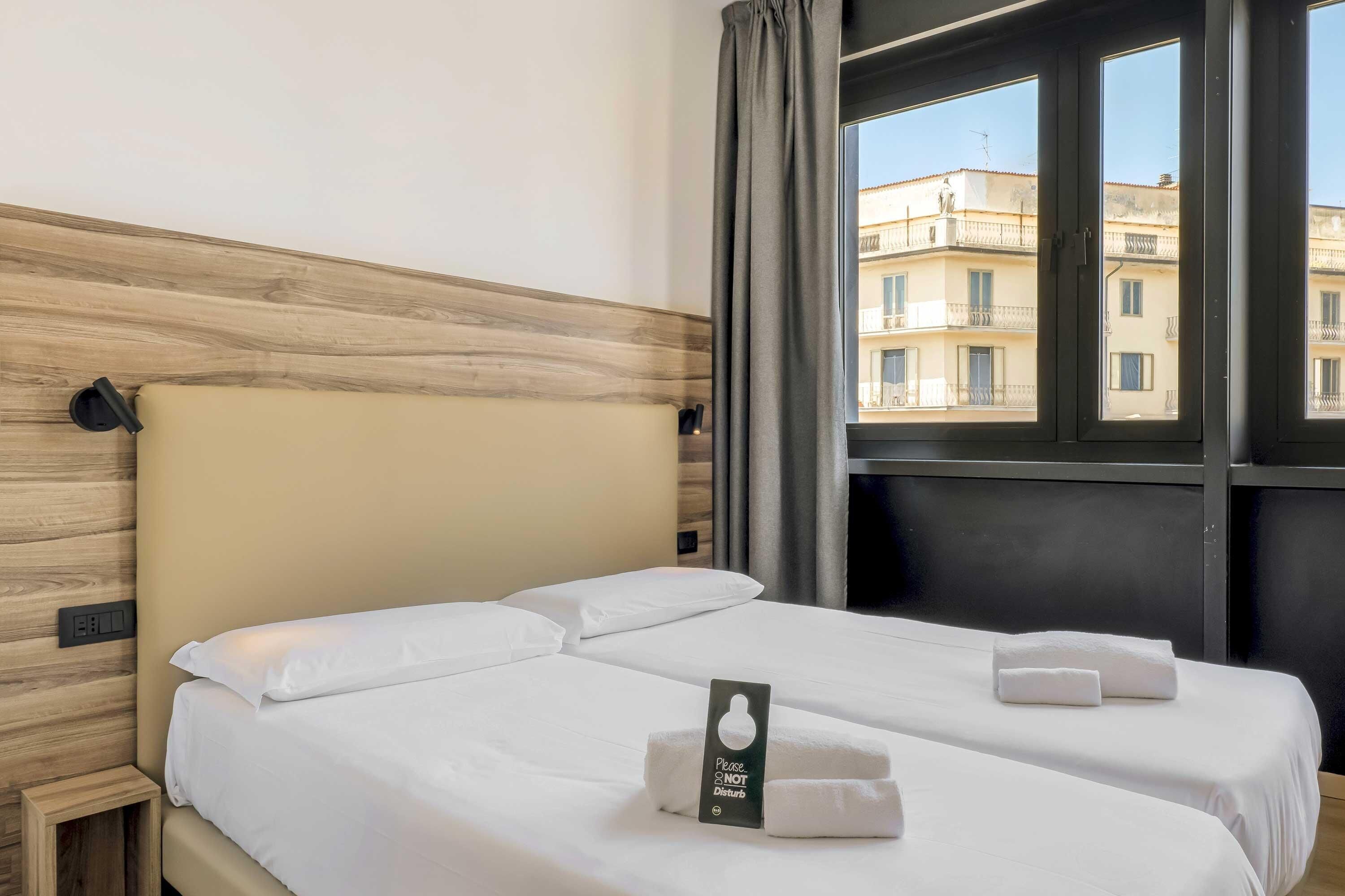 Habitación de hotel con dos camas, cabecero de madera, ventana con cortinas y un cartel en la cama.