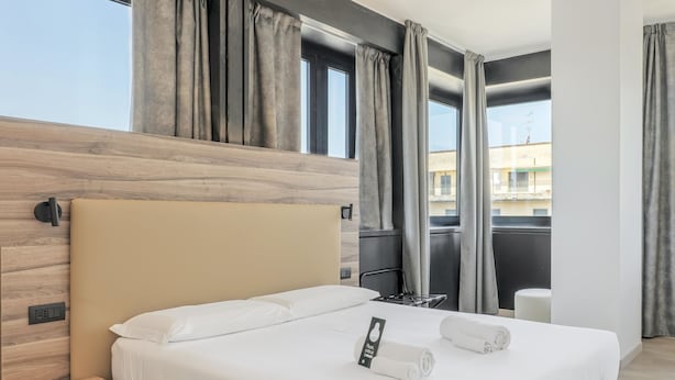 A modern hotel room with a large bed, a wooden headboard, and a view of the city through large windows. - B&B Hotel Prato City Center (Prato)